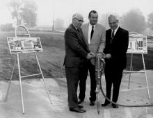 Three men in suits holding onto a jackhammer with images of a building on easels behind them