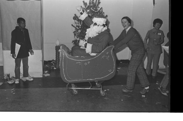 Black and white photograph. A black man dressed as Santa Clause is sitting in a makeshift sleigh made out of a shopping cart, cardboard and decorated with glitter. His fake beard and the trim on his hat, sleeves and belt are made out of cotton balls. A smiling woman in a white turtleneck, cardigan, and grid pants is pushing the sleigh. A black child is standing on the far left, frowning and with a bundle of white paper tucked under his arm. Another woman in a suit is in the background. Behind Santa is a decorated tree and giant plastic candle. Debris of confetti litters the floor.
