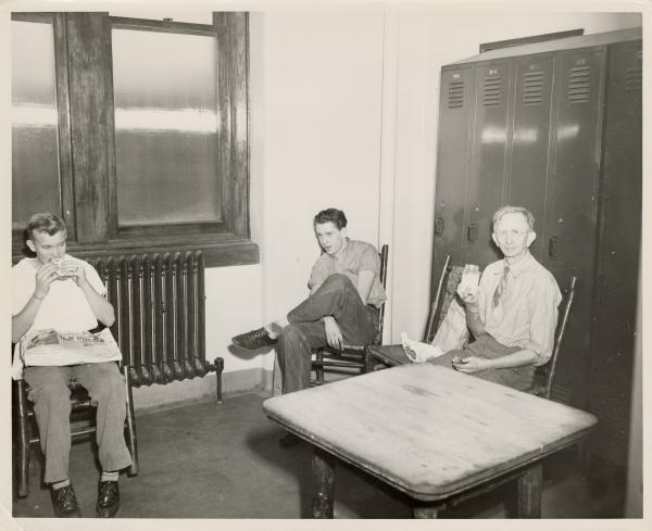 Black and white photograph. Three men, two younger and one older, sit on wooden chairs in a locker room. The young man on the far left is eating a hamburger and has a newspaper on his lap. The older man is sitting on the far right and has glasses and wearing a tie. His sleeves are rolled up to the elbow. He is looking into the camera and is holding up a mason jar filled with milk. Between them is the last young man, who has his legs crossed and is looking into the distance, away from the camera. Behind all of the men is a radiator and a window.