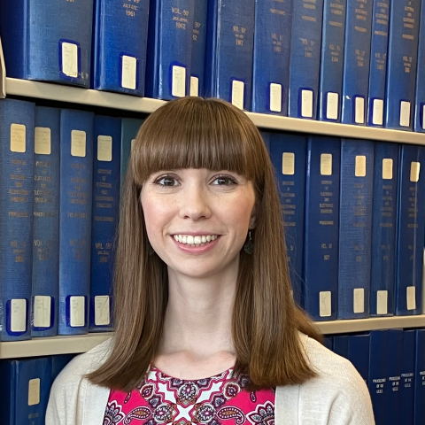 White woman with long brown hair and bangs. Smiling in front of a bookshelf.